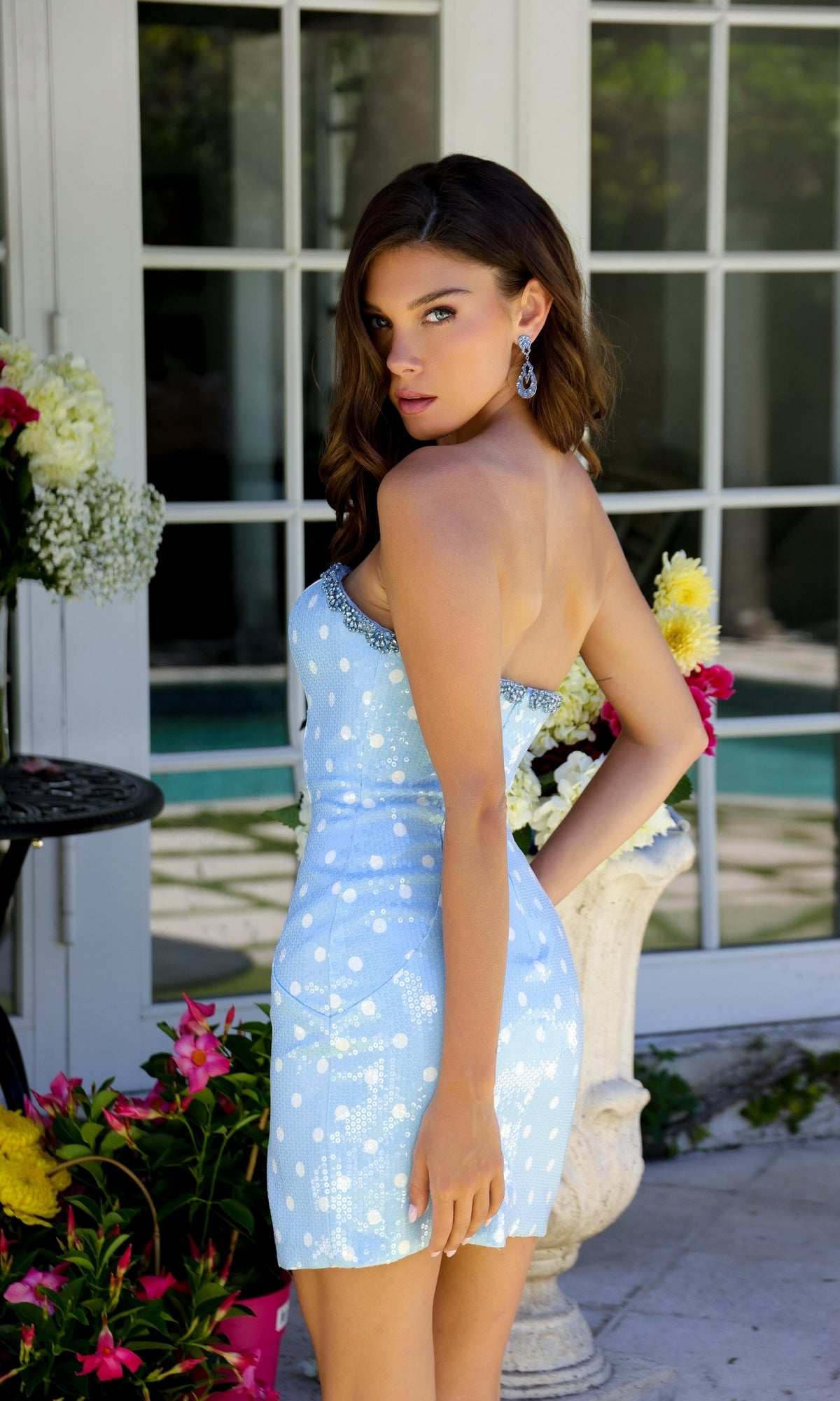 A woman wearing the Ava Presley 29159 Polka-Dot Sequin Homecoming Dress stands outdoors near a window and flowers, looking over her shoulder at the camera.